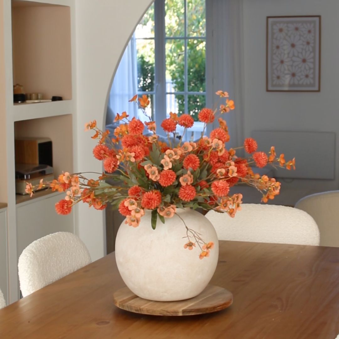 Vase of orange flowers on a wooden table in a bright room.