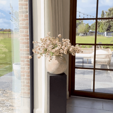 Vase with flowers on a stand by window overlooking a garden and patio.