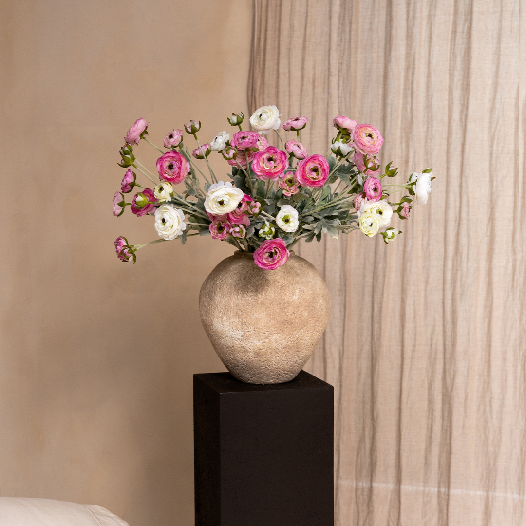 A textured tan vase of pink and white flowers sits on a black pedestal against a neutral background.