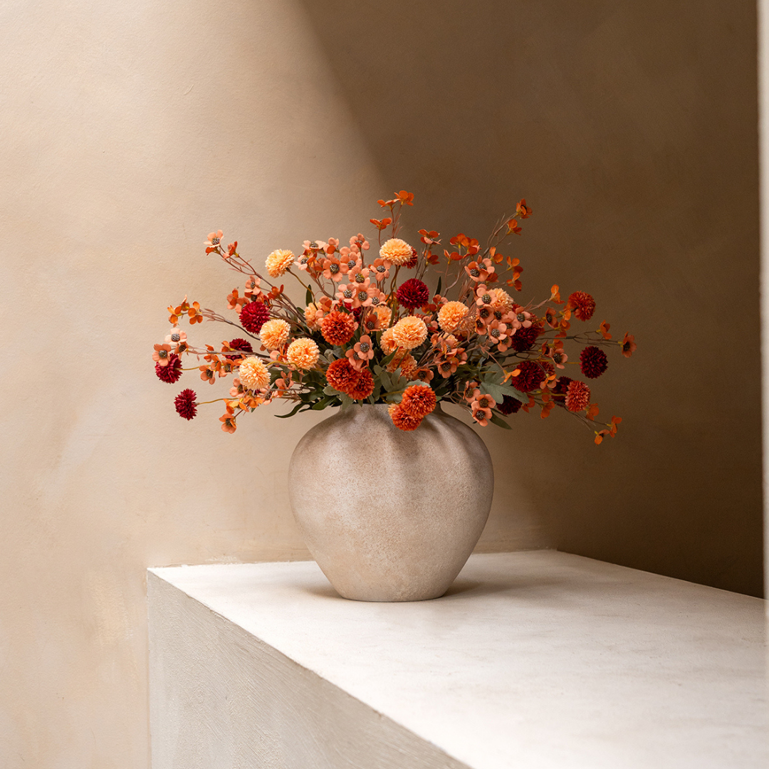 A textured vase with an arrangement of red and orange flowers sits on a white shelf against a wall.