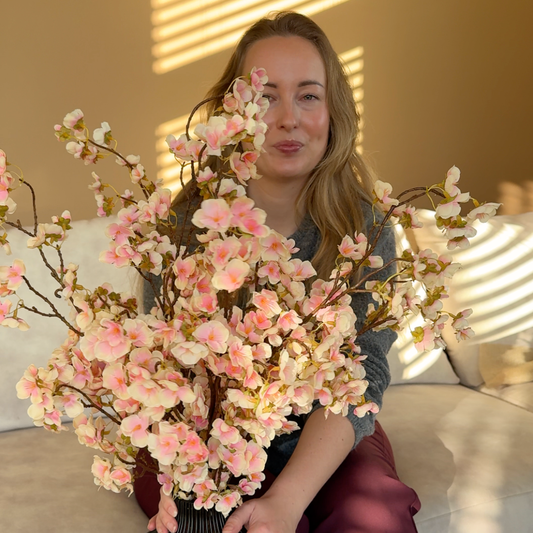 A woman with long blonde hair sits on a couch holding a large vase of pink cherry blossoms.
