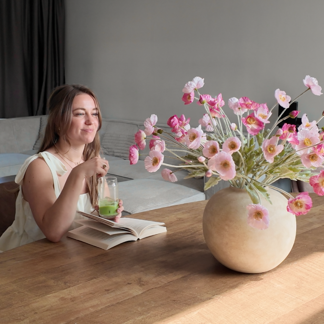 A woman sits at a wooden table with a book, a green drink, and a large vase of pink flowers.