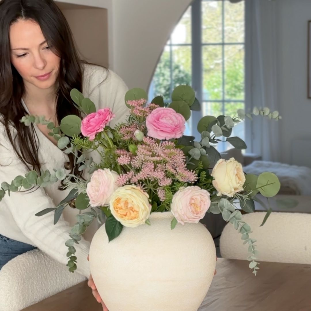 A woman in a white sweater arranges a large bouquet of pink and cream flowers in a white vase.