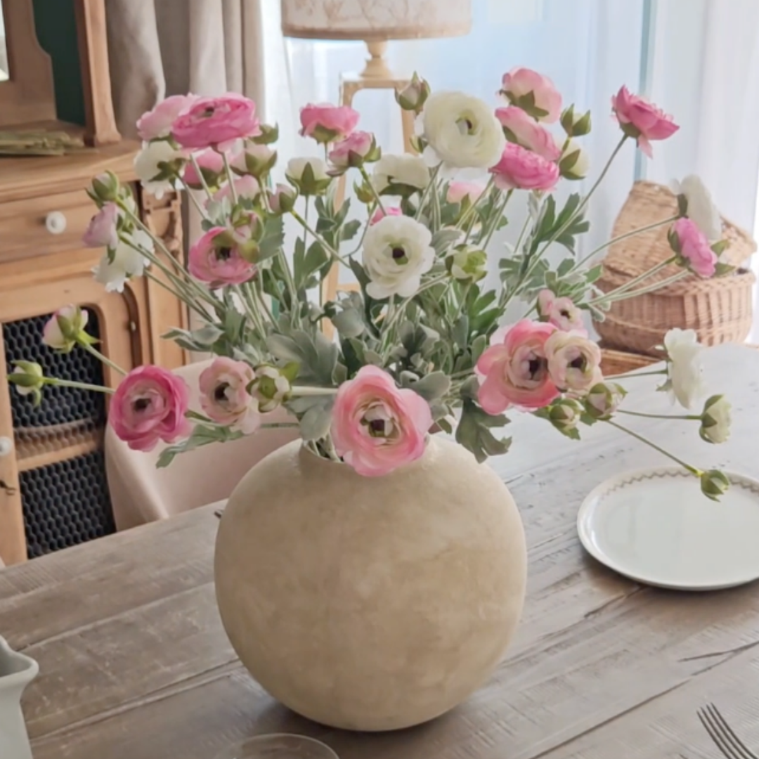 A bouquet of pink and white ranunculus flowers in a round, light-colored vase on a wooden table.