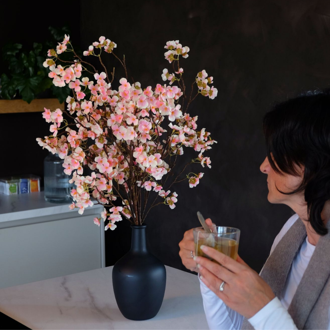 A woman holding a cup, sitting by a vase of pink flowers.