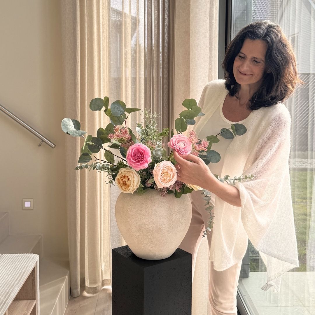 A woman with dark hair arranges a large bouquet of roses and eucalyptus in a stone vase.