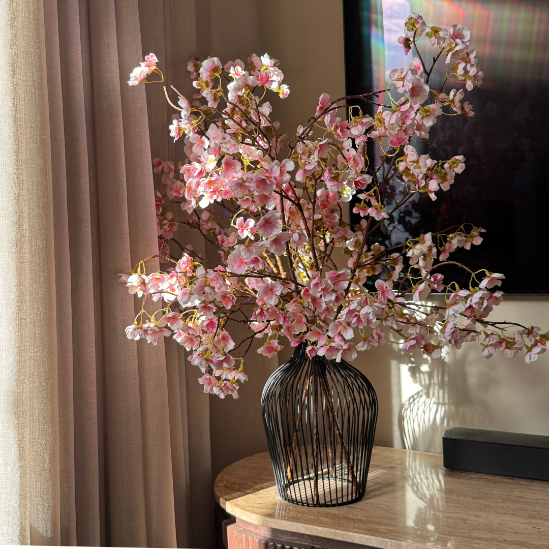 Pink cherry blossom branches in a black wire vase sit on a sunlit marble table next to a curtain.