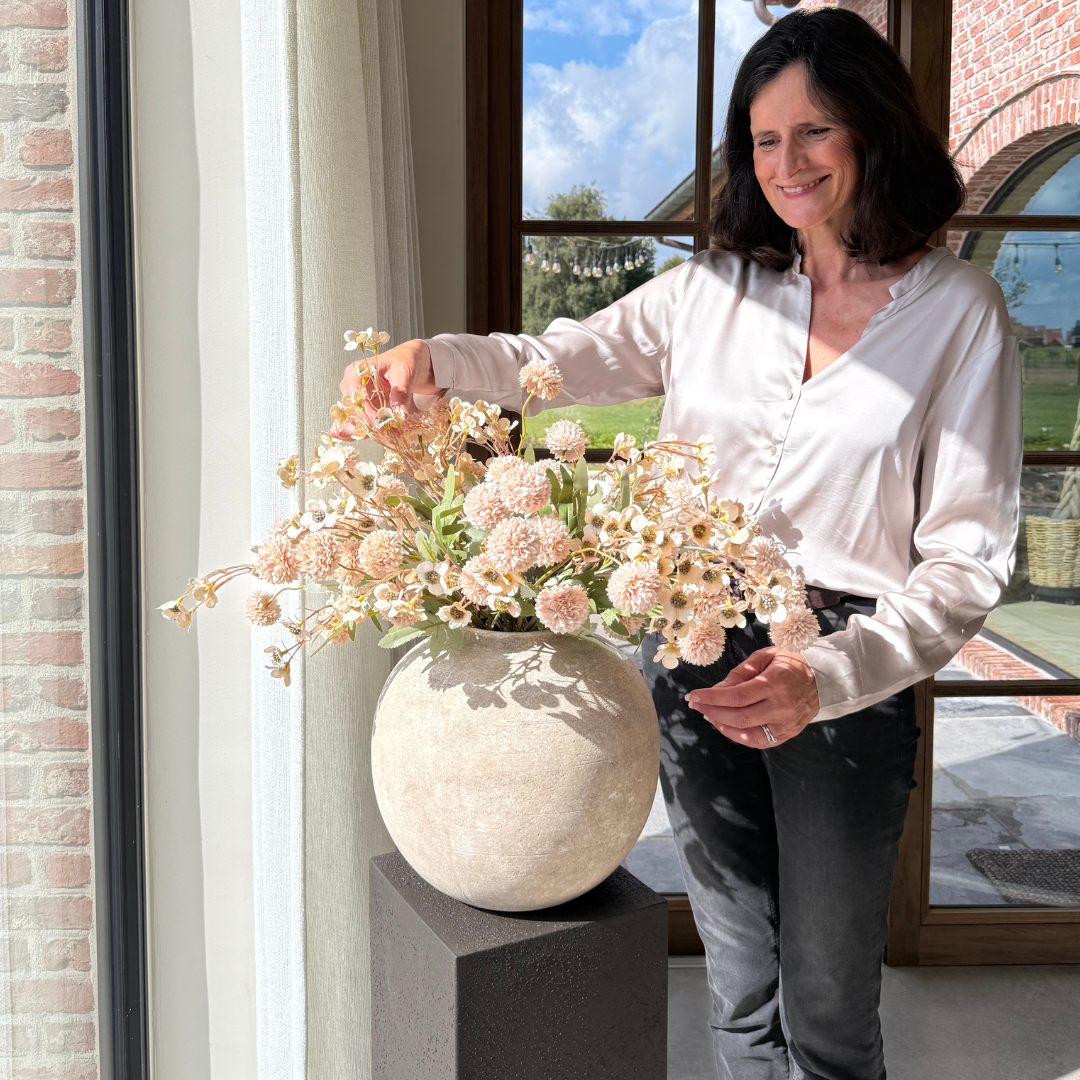 Woman arranging flowers in a round vase by a sunny window.