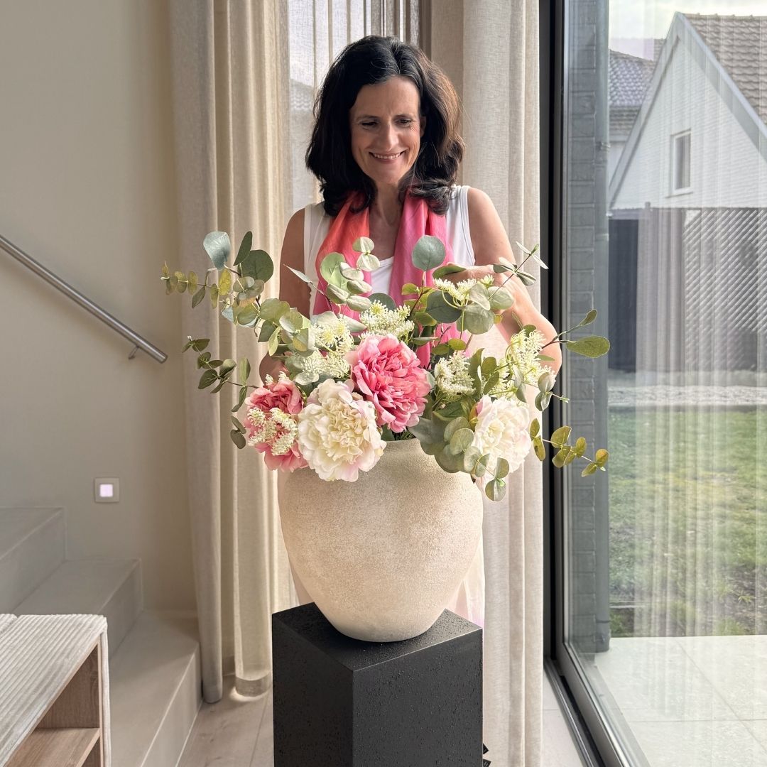 A woman smiles as she arranges a large bouquet of pink and white flowers in a vase.
