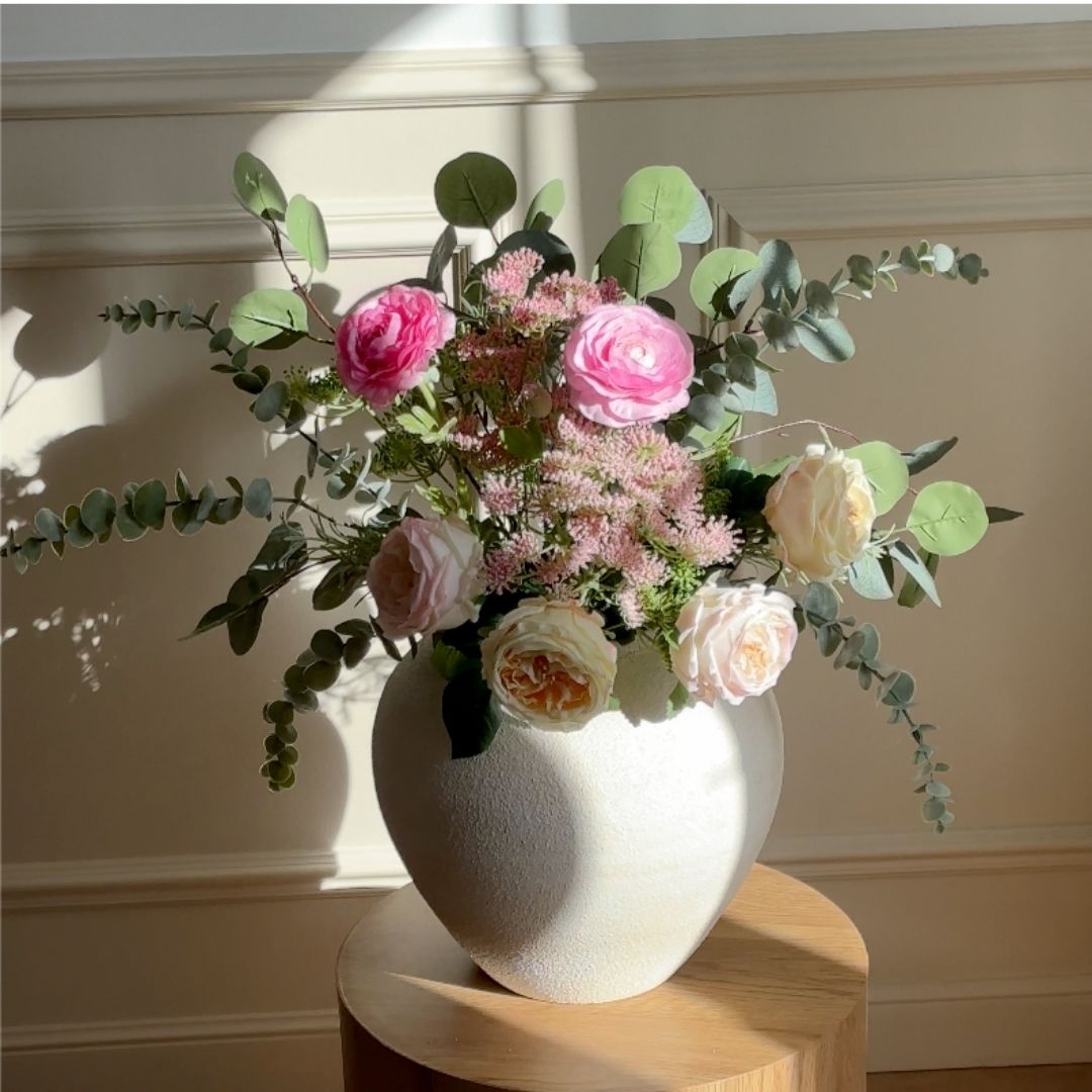A textured white vase holds an arrangement of pink and cream roses and eucalyptus leaves on a wooden table.