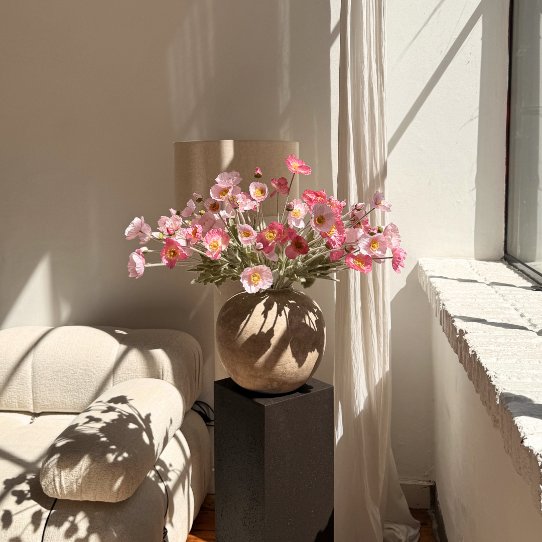 A sunlit room with a round vase of pink poppies on a dark pedestal next to a chair.