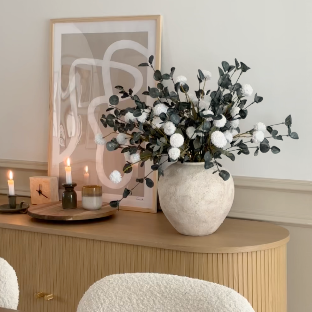 Vase with white flowers and greenery on sideboard next to abstract art and candles.