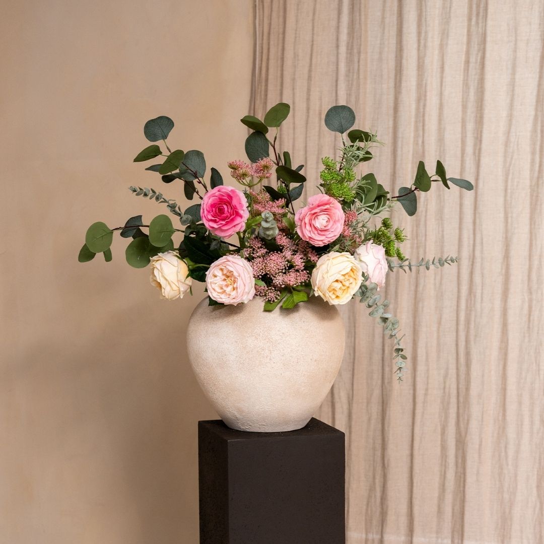 A floral arrangement of pink and cream roses and eucalyptus in a textured vase on a dark pedestal.
