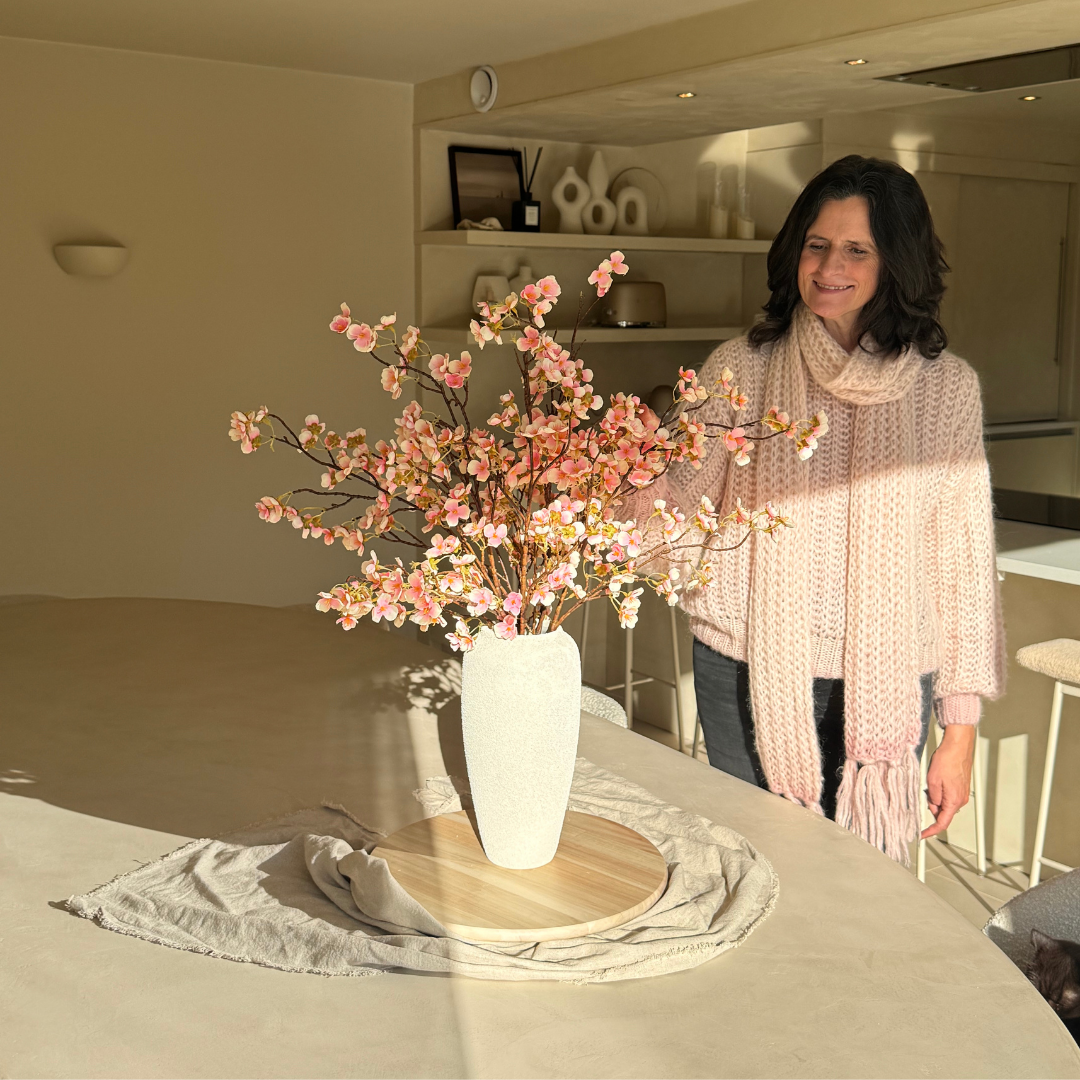A woman in a cozy sweater arranges pink flowers in a vase on a round table.