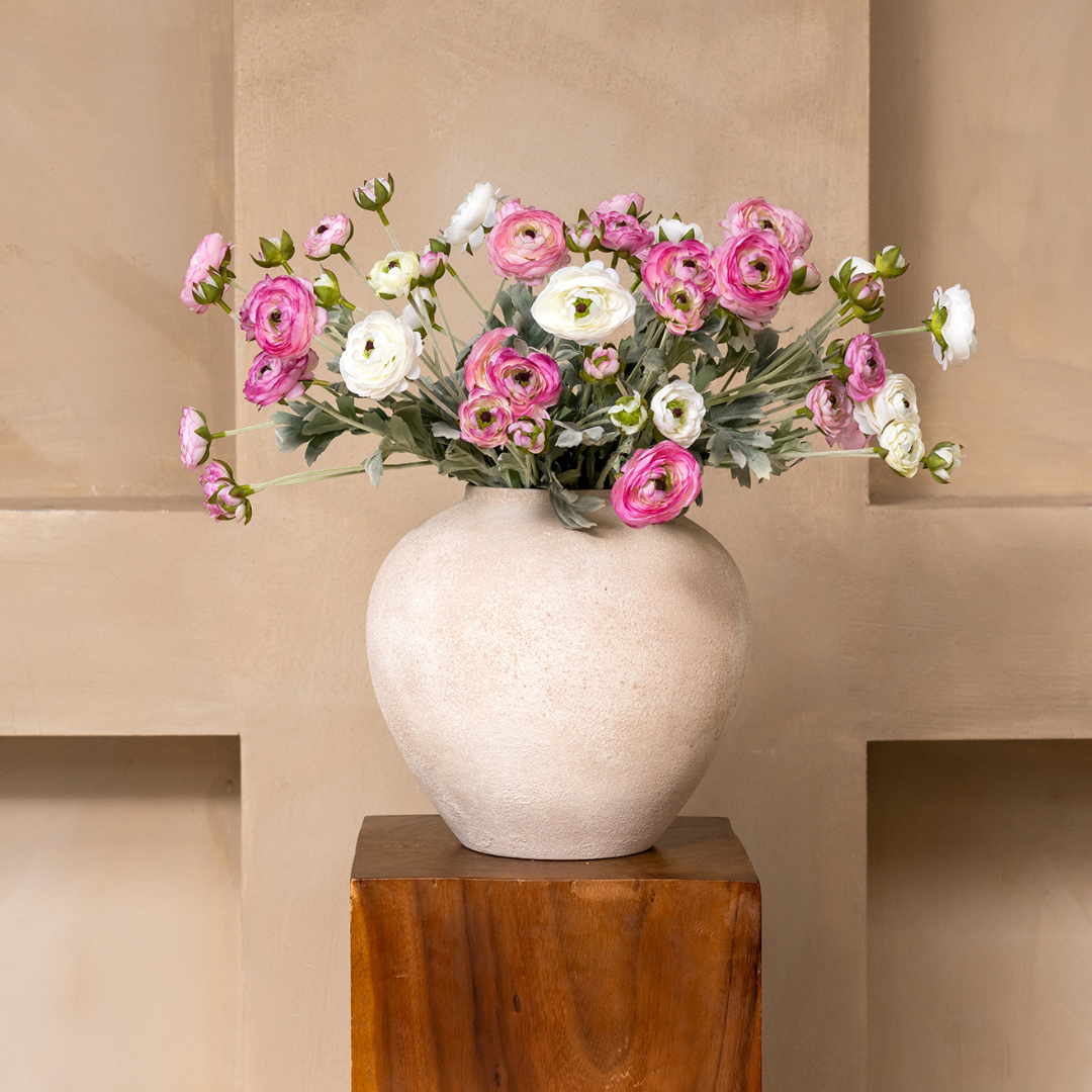 A bouquet of pink and white ranunculus flowers in a textured beige vase on a wooden block.