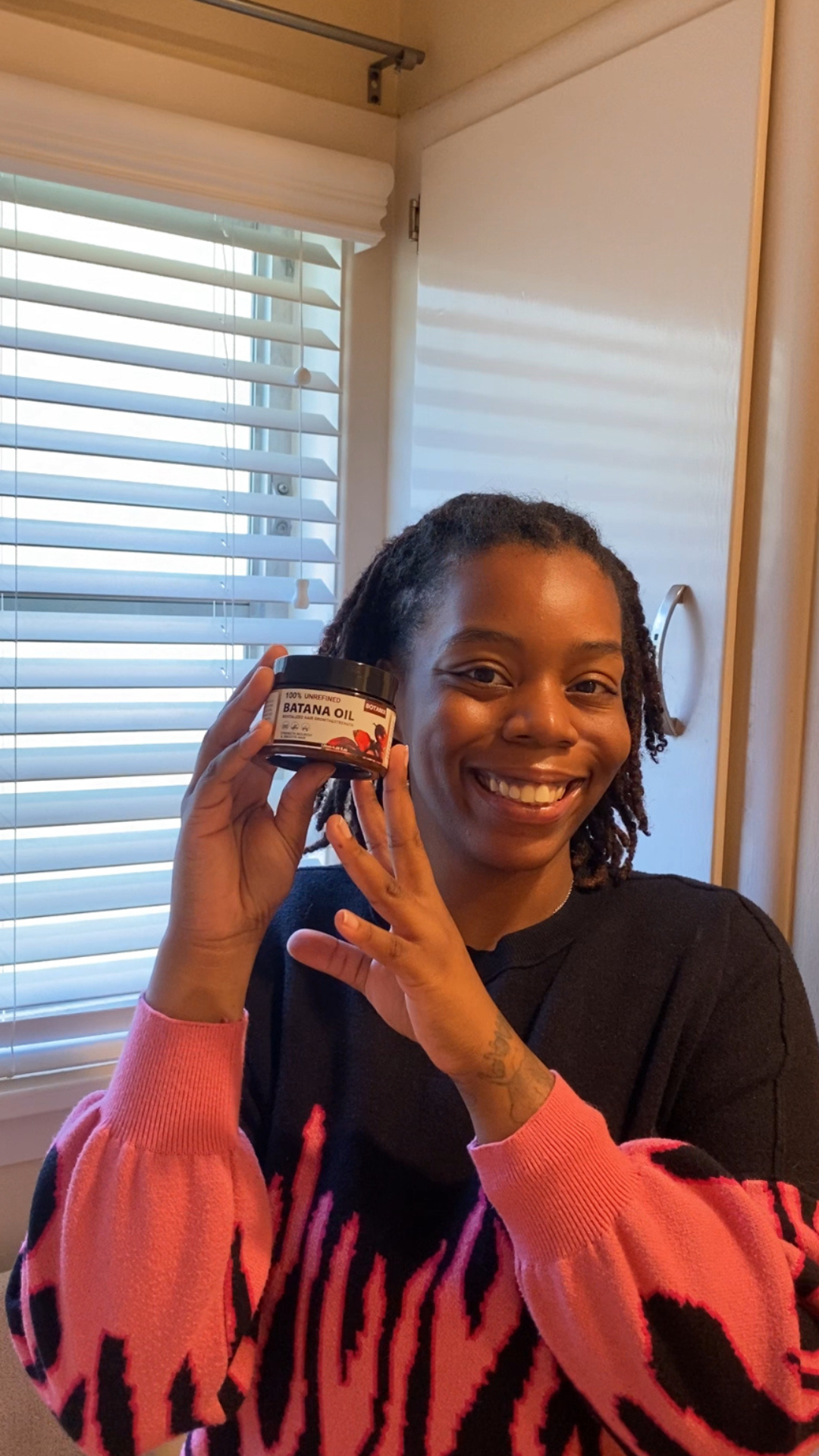 Person smiling and holding a container of Batana oil indoors.