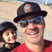 A smiling man in a baseball cap and sunglasses takes a selfie with a child wearing a helmet.