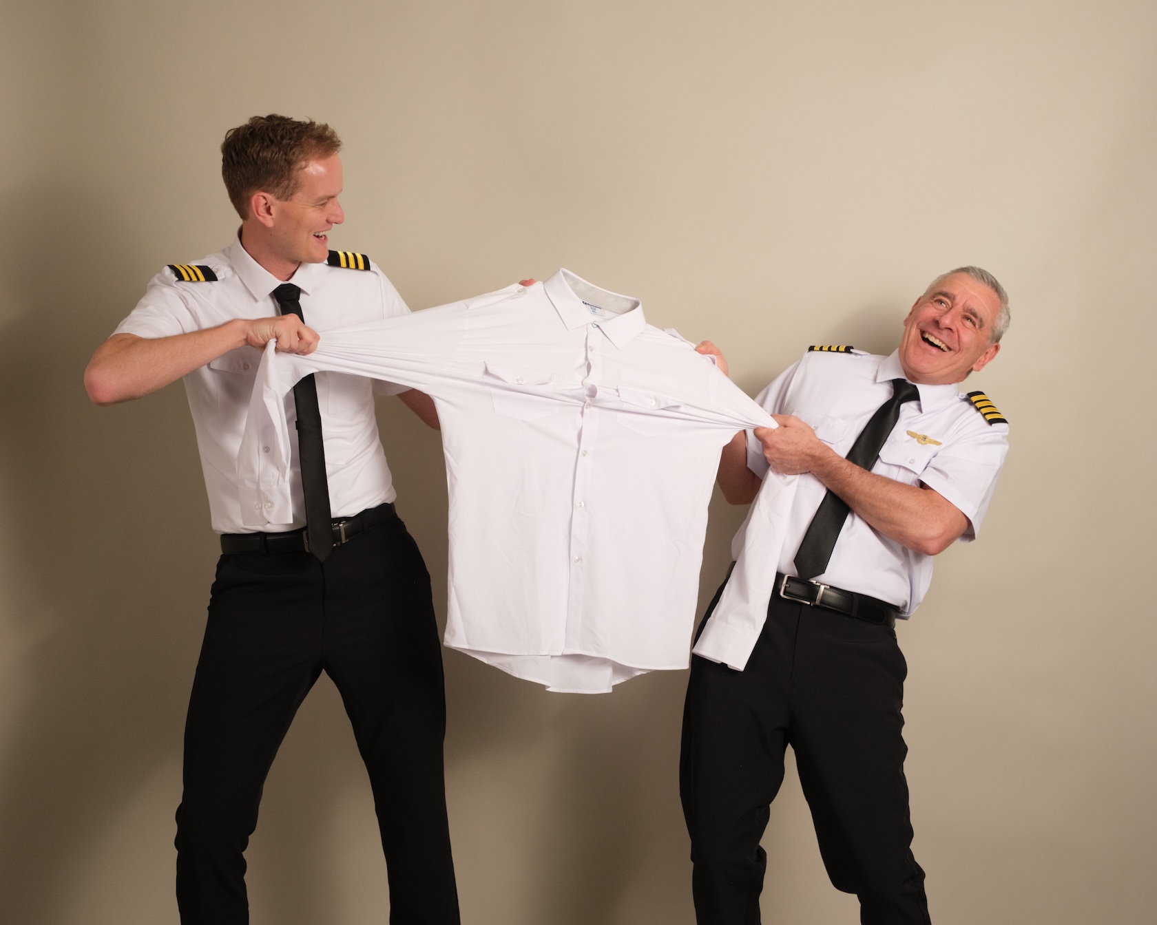 Two smiling men dressed as pilots stretch a white button-down shirt between them against a plain background.