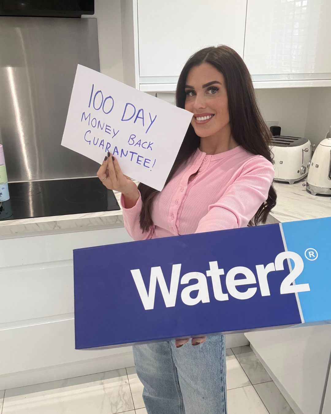 A woman in a kitchen holds a Water2 box and a sign that says '100 Day Money Back Guarantee!'