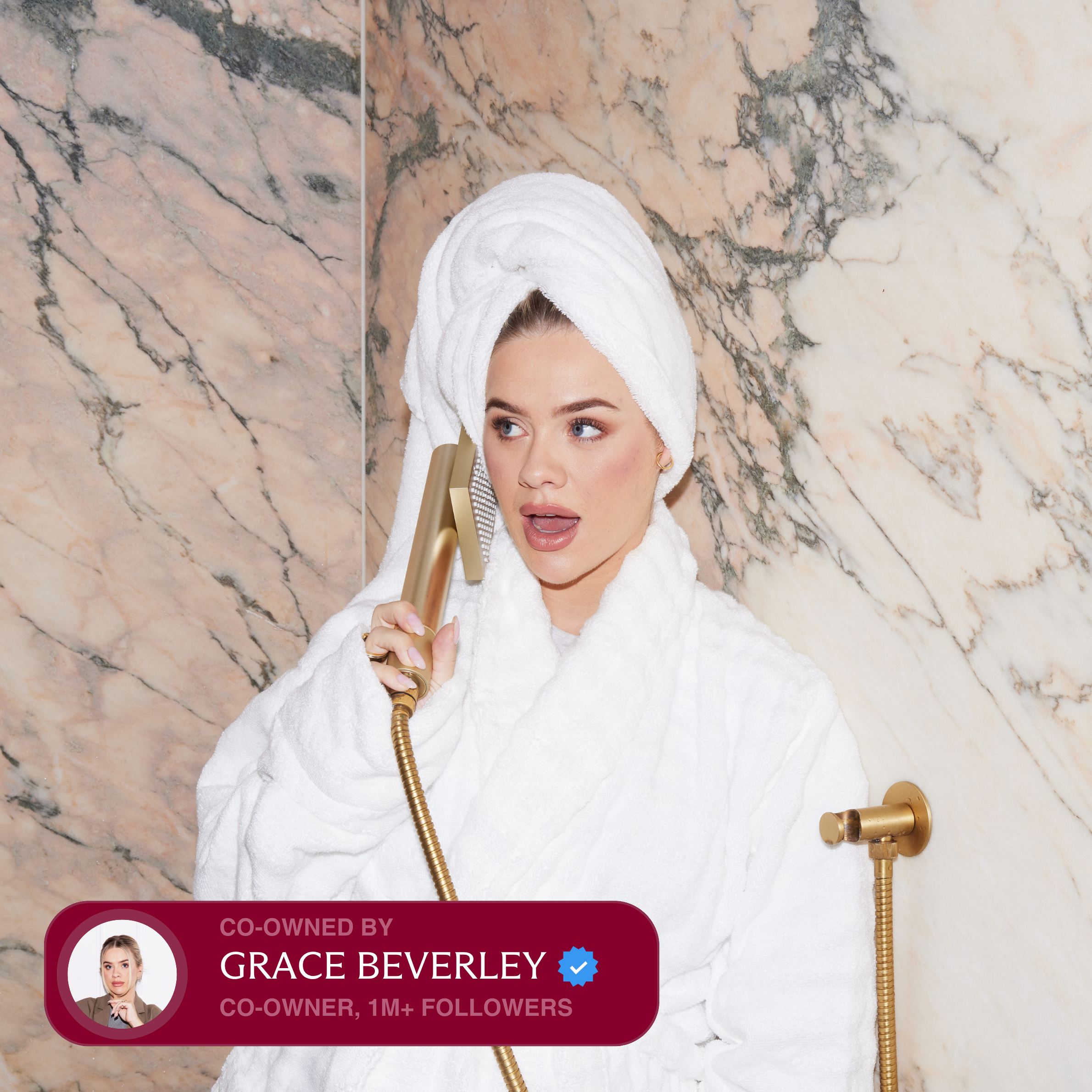 A woman in a bathrobe and hair towel holds a gold shower head to her ear like a phone.