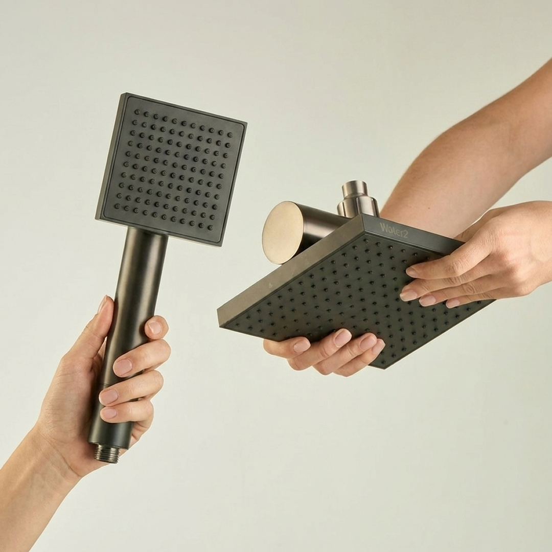 Hands holding a dark, square handheld shower wand and a matching rain shower head against a white background.