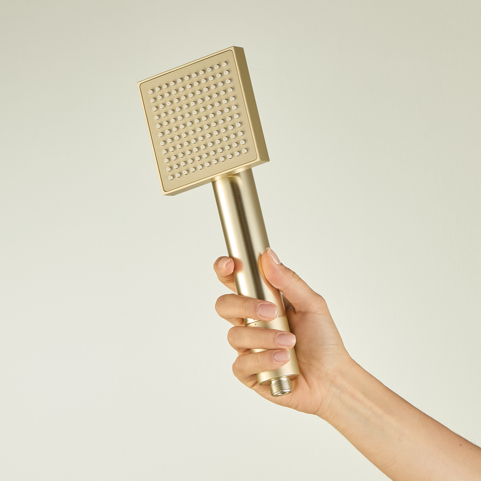 A hand holding a square, gold-colored handheld showerhead against a plain background.