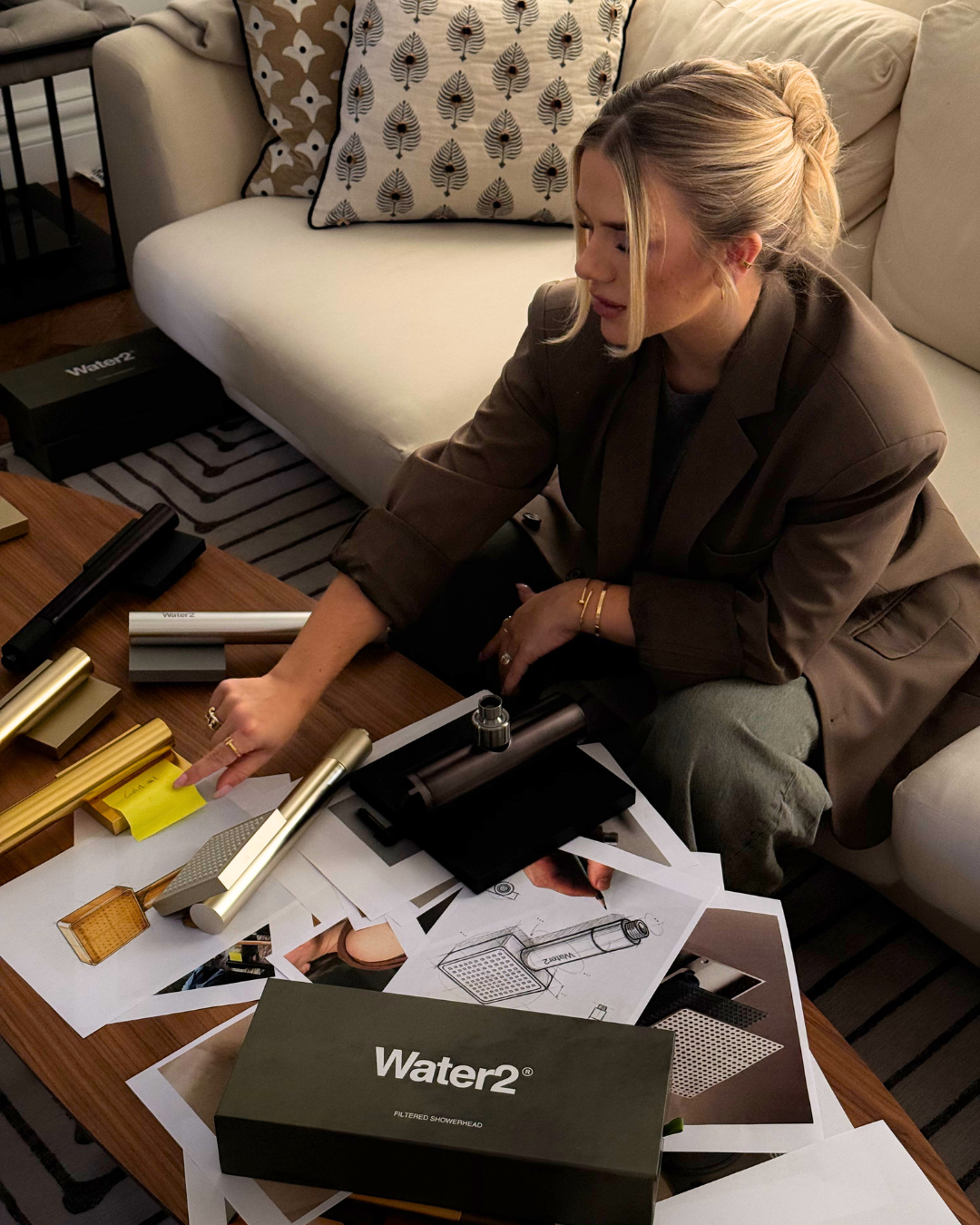 A woman reviews showerhead product designs, material samples, and packaging spread across a coffee table.