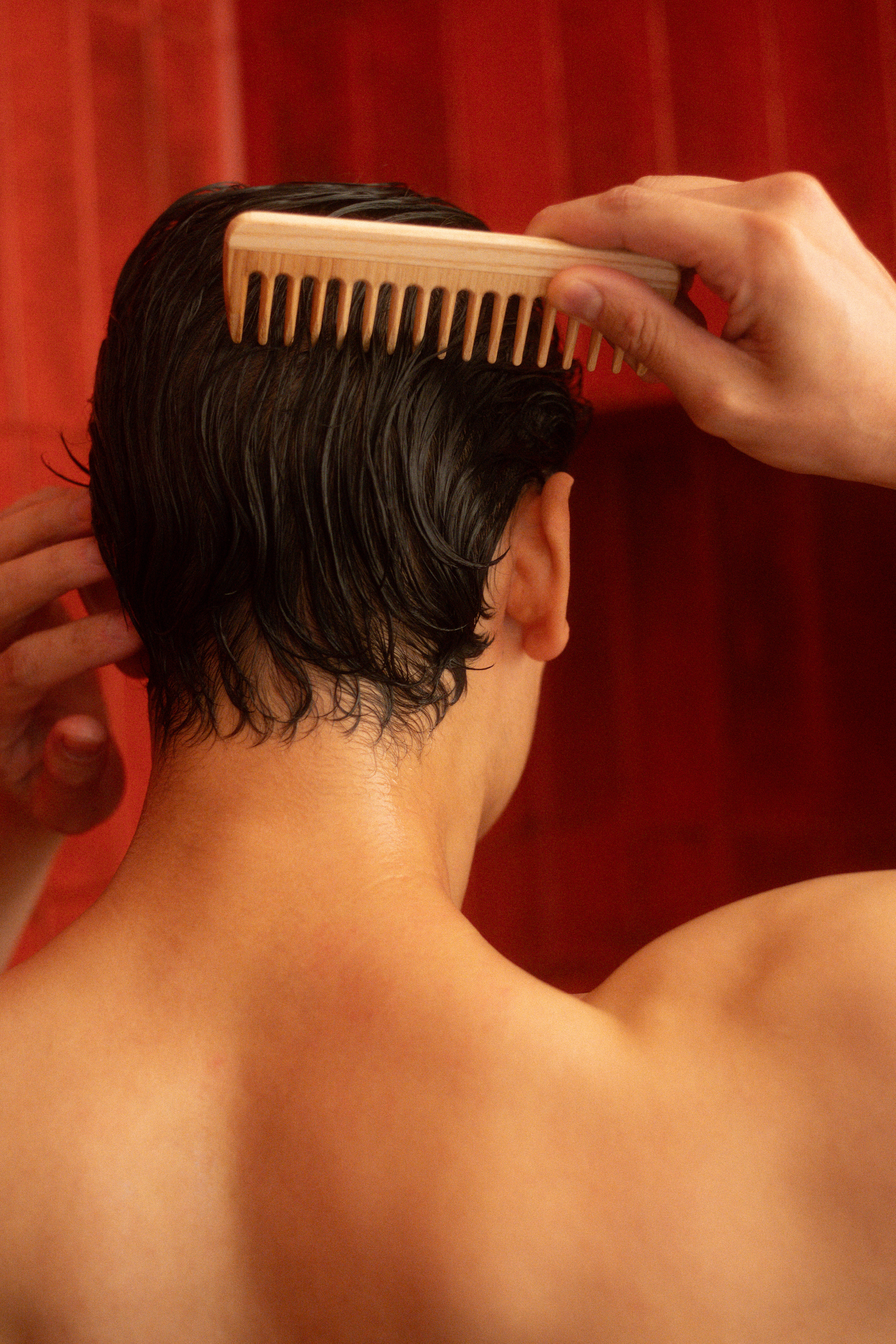 A person viewed from behind combing their wet, dark hair with a wooden comb against a red background.
