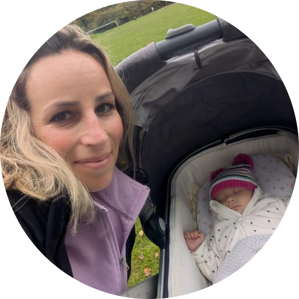 A woman takes a selfie with a baby sleeping in a pram outdoors on a grassy field.