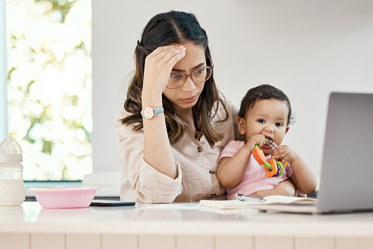 A tired-looking mother holds a baby in her lap while sitting at a desk with a laptop.
