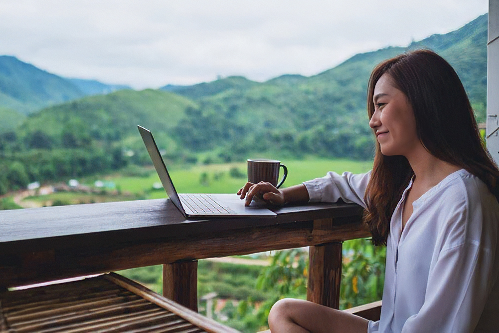 A woman sits with a laptop and a coffee mug on a balcony overlooking lush green mountains.