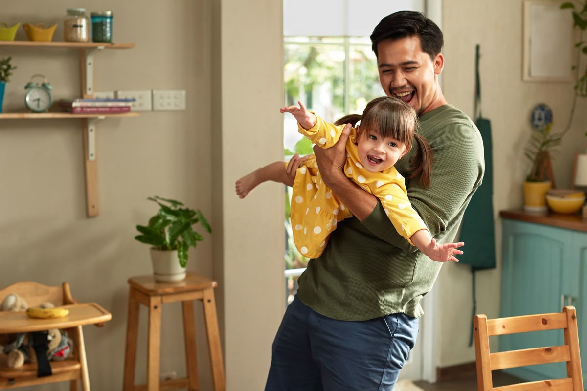 A smiling man playfully lifts a laughing toddler in a yellow polka-dot dress inside a home.
