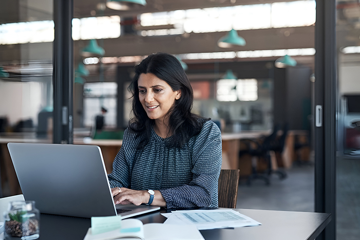 A woman with dark hair sits at a desk in an office, smiling while using a laptop.
