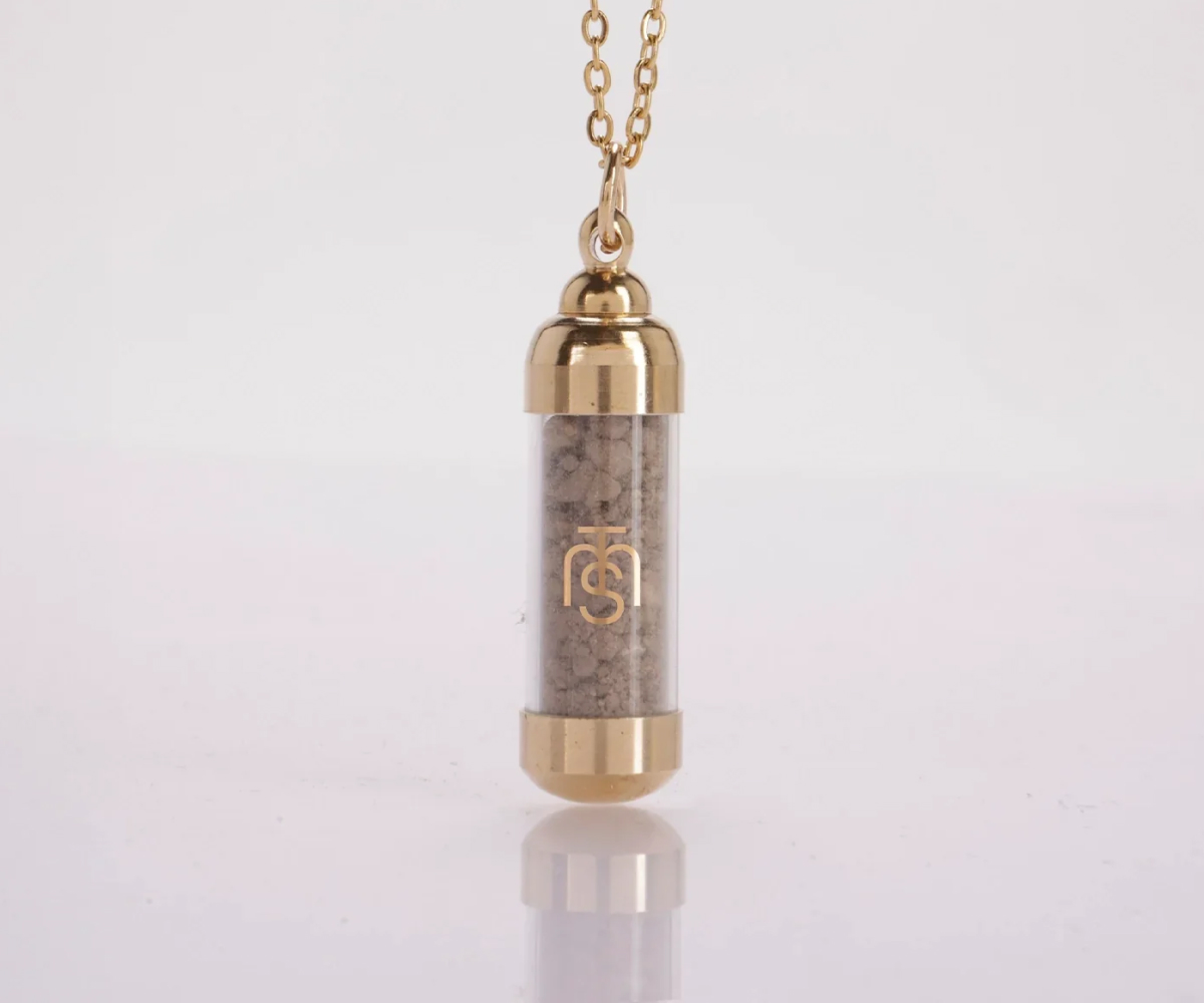 A gold necklace with a clear vial pendant containing a grey, sand-like substance on a white surface.