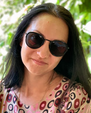 A person smiling outdoors with greenery in the background.