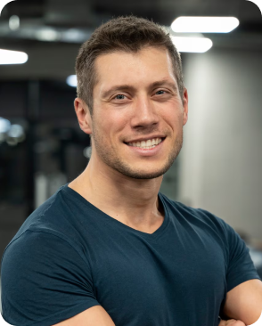 Smiling person in a gym setting with arms crossed, wearing a blue shirt.