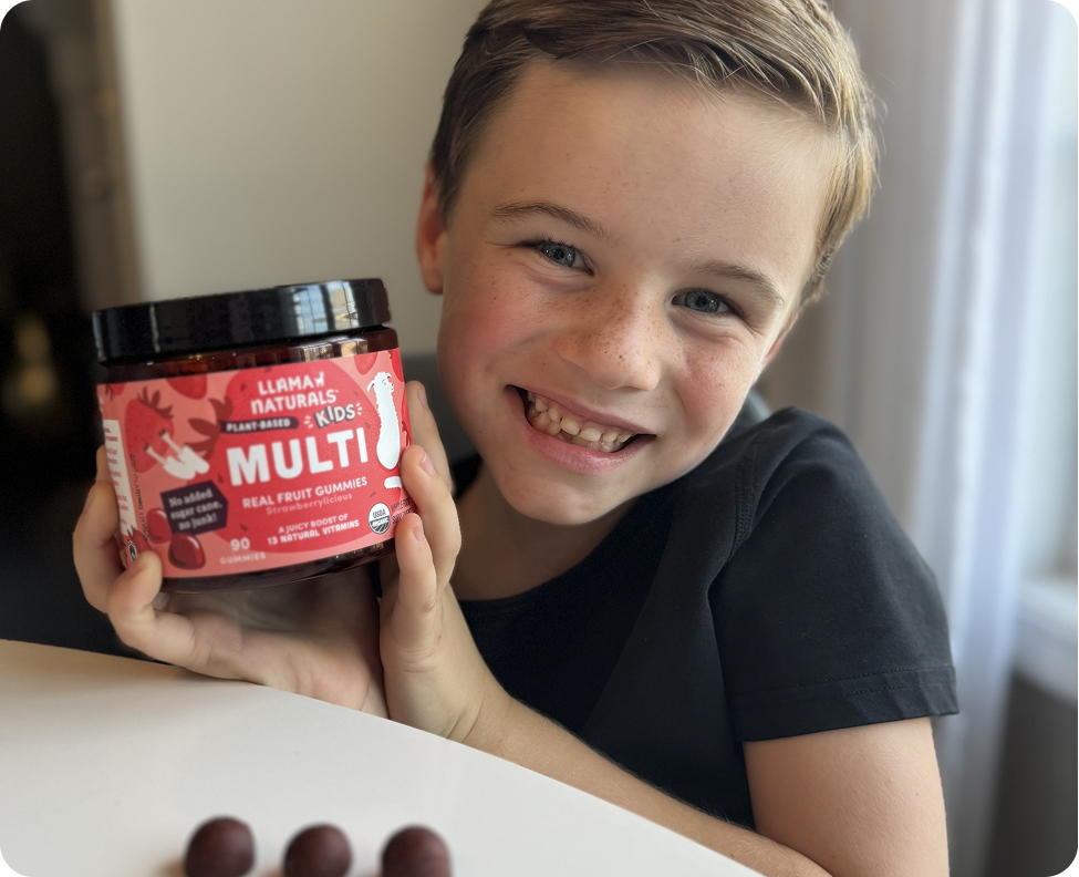 A smiling young boy in a black shirt holds up a jar of Llama Naturals Kids Multi gummies.
