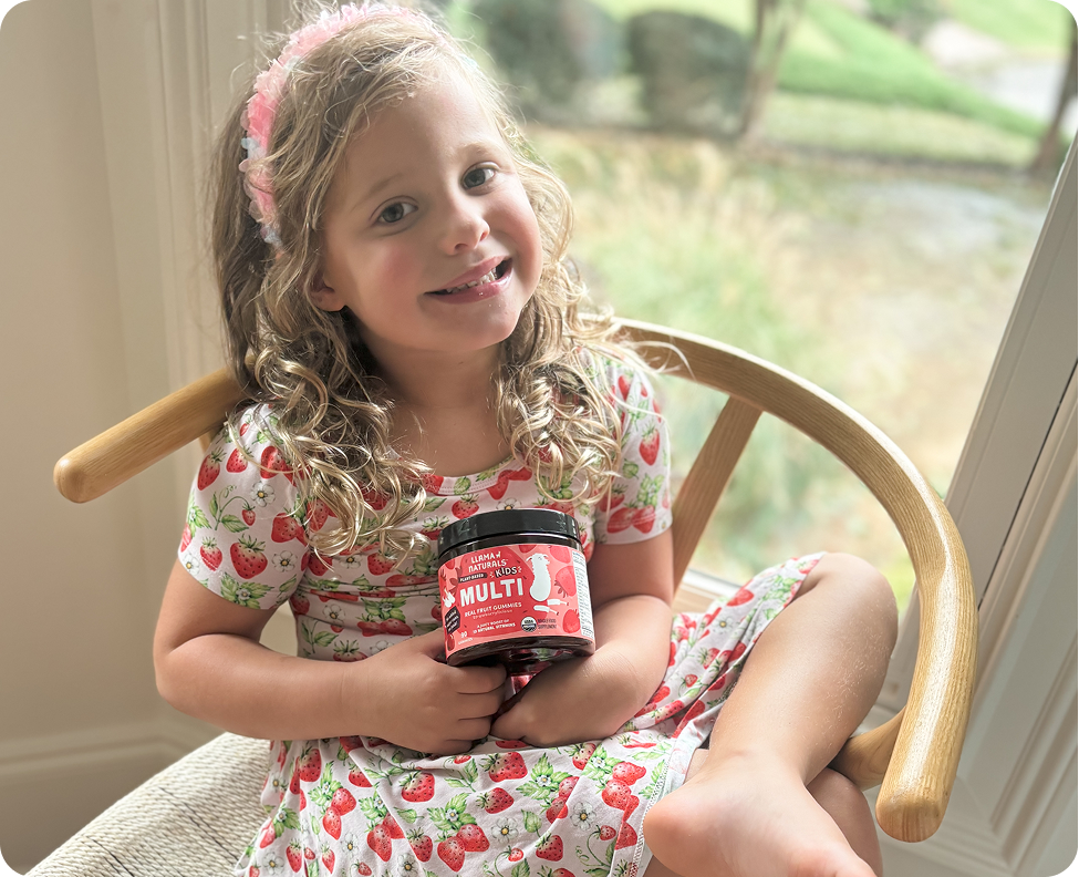 A smiling young girl in a strawberry dress holds a jar of Llama Naturals Kids Multi gummies.