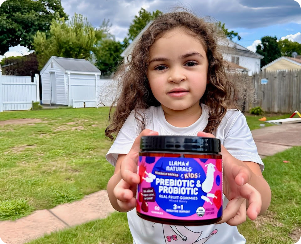 A young girl stands in a backyard, holding a jar of Llama Naturals kids prebiotic and probiotic gummies.
