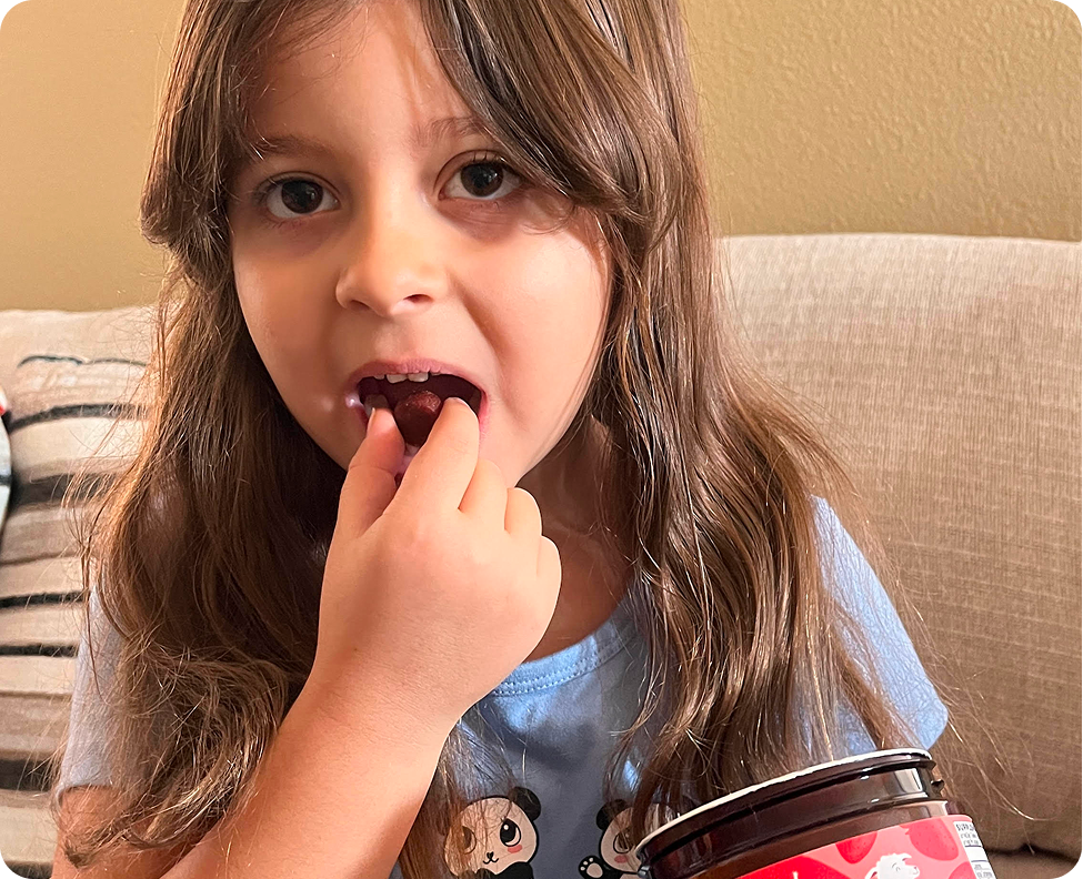A young girl with long brown hair looks at the camera while putting a small, round food item in her mouth.