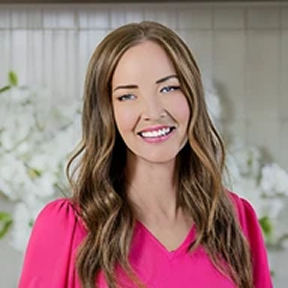 A headshot of a smiling woman with long brown hair and blue eyes, wearing a pink top.