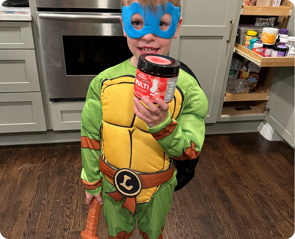 A young boy in a Teenage Mutant Ninja Turtle costume holds a jar of children's multivitamins.