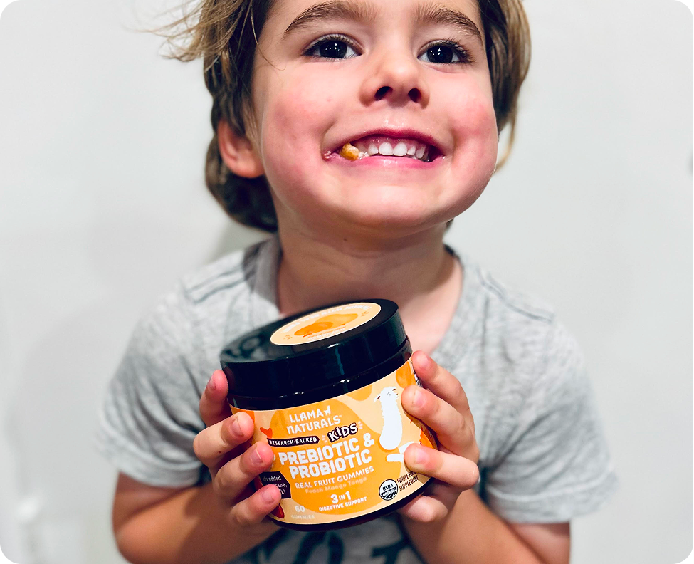 A smiling boy holds a jar of Llama Naturals Kids Prebiotic & Probiotic gummies with one in his mouth.