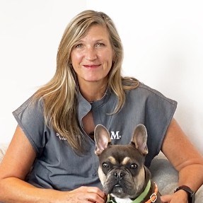 A woman with blonde hair in a grey top sits next to a French bulldog.