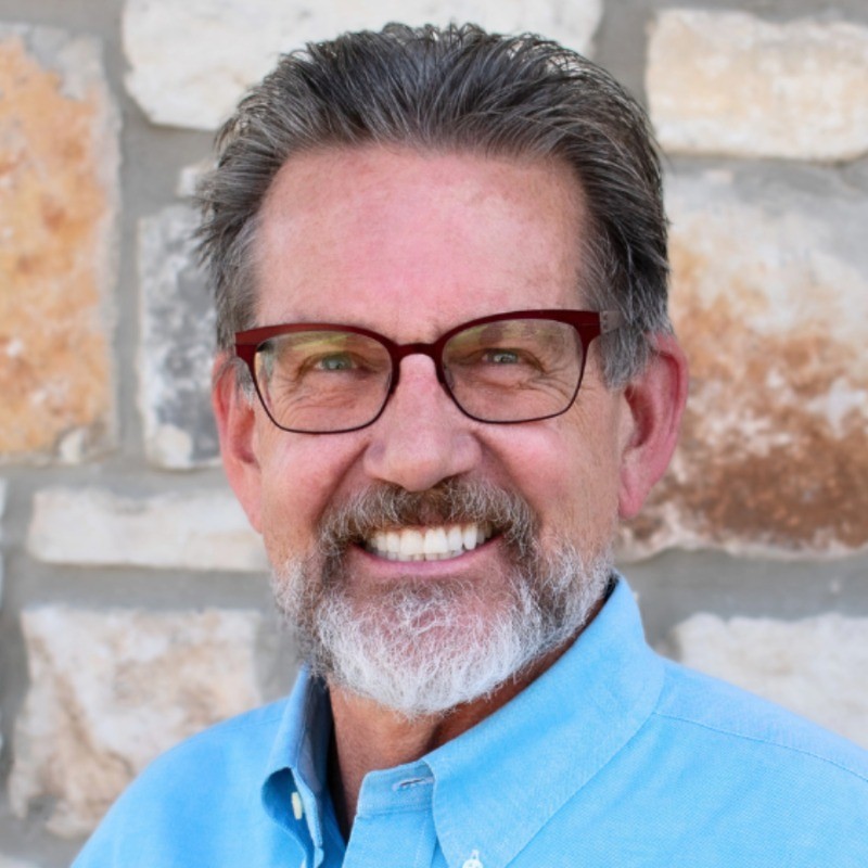 A smiling man with glasses, a gray beard, and a light blue shirt standing in front of a stone wall.