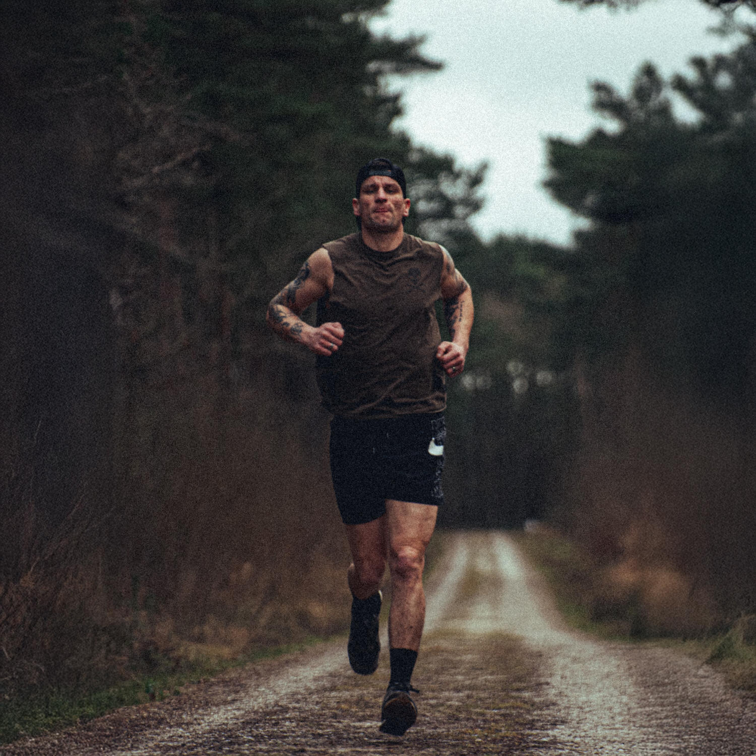 Man running on a forest trail wearing a cap and sleeveless shirt.