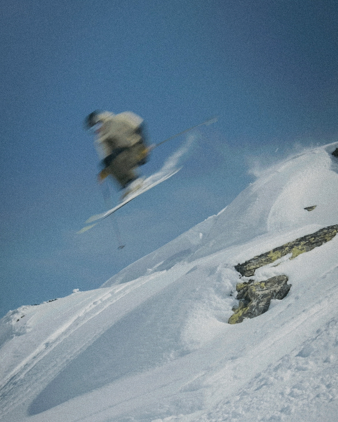 A motion-blurred shot of a skier jumping off a snowy mountain against a clear blue sky.