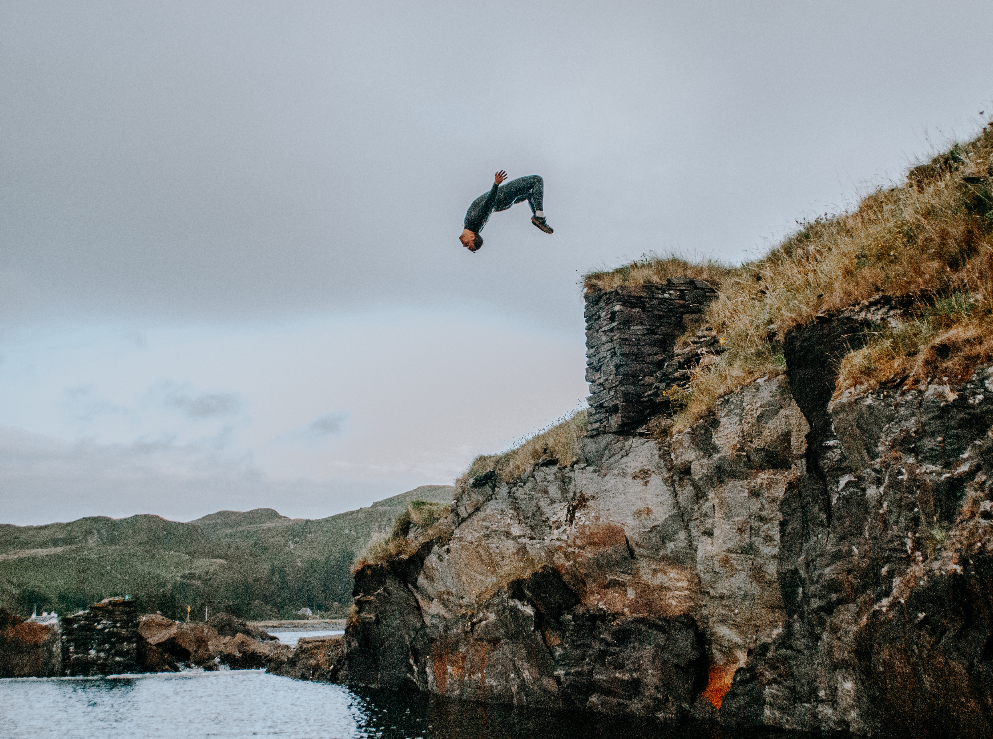 A person does a backflip off a rocky, grass-topped cliff into the water below under an overcast sky.