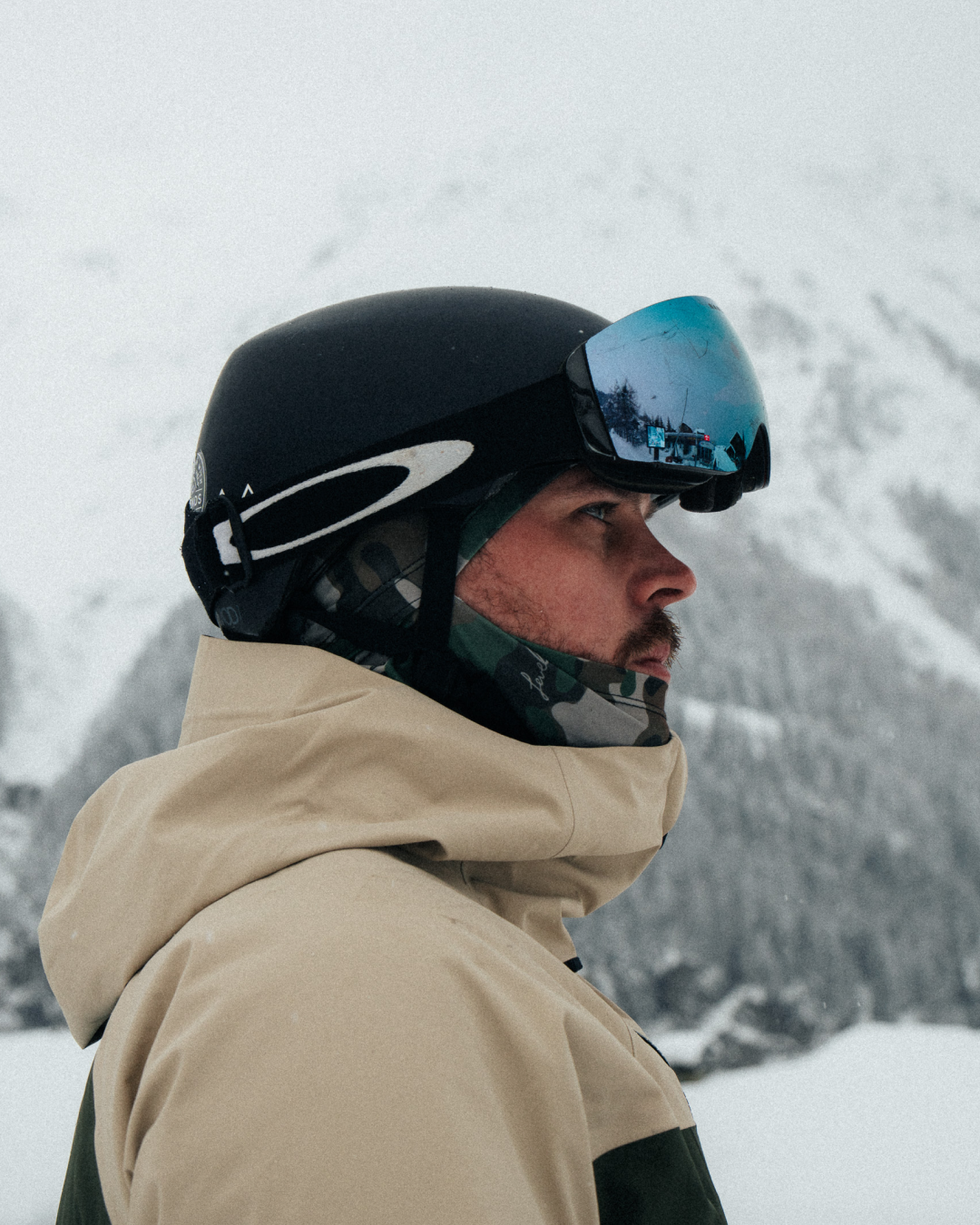 A person in a ski helmet and goggles looks to the side against a snowy mountain background.
