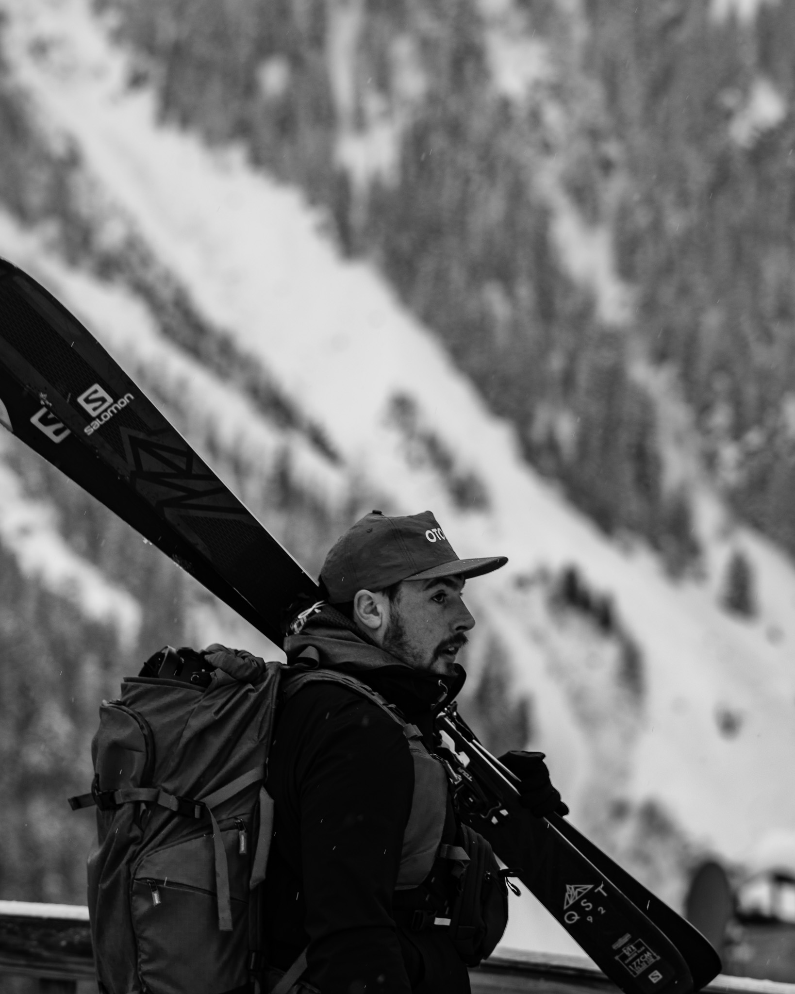 A man in a cap with a backpack carries skis on his shoulder in a snowy landscape.